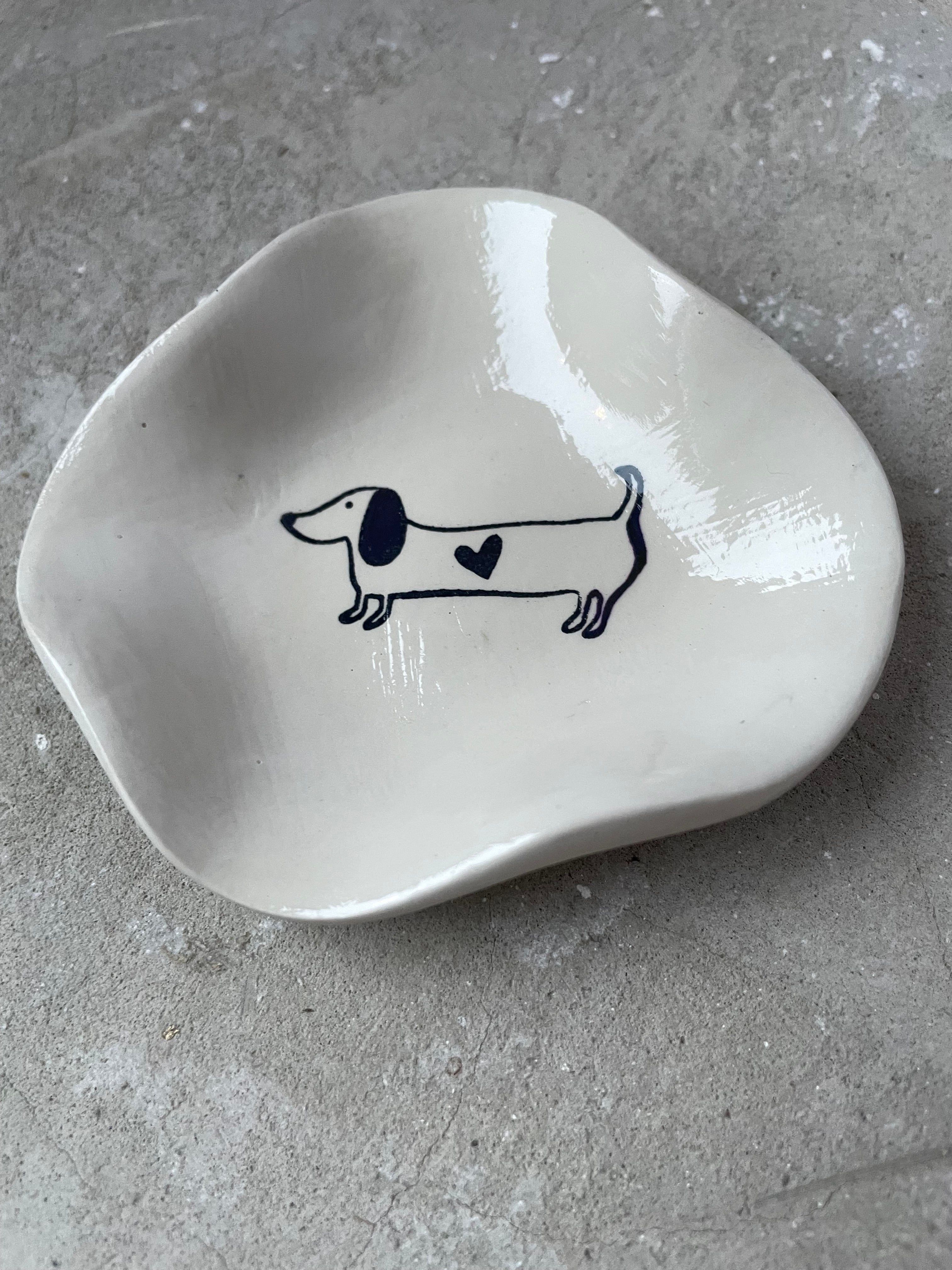 Gegossenes Keramikschälchen in warmem Pantone-Beige mit High-Gloss-Oberfläche. Innen ein gezeichneter Dackel mit Herz – schlicht, modern und super für Tierfreunde. Perfekt als Schmuckschale, Ablage für Schlüssel oder kleine Lieblingsstücke. Handmade, einzeln gegossen, geschliffen und veredelt. Langlebig, pflegeleicht und ein süßes kleines Wohnaccessoire für Bad, Schlafzimmer oder Schreibtisch. Schöne Geschenkidee für Hundeliebhaber und Dackelfans.