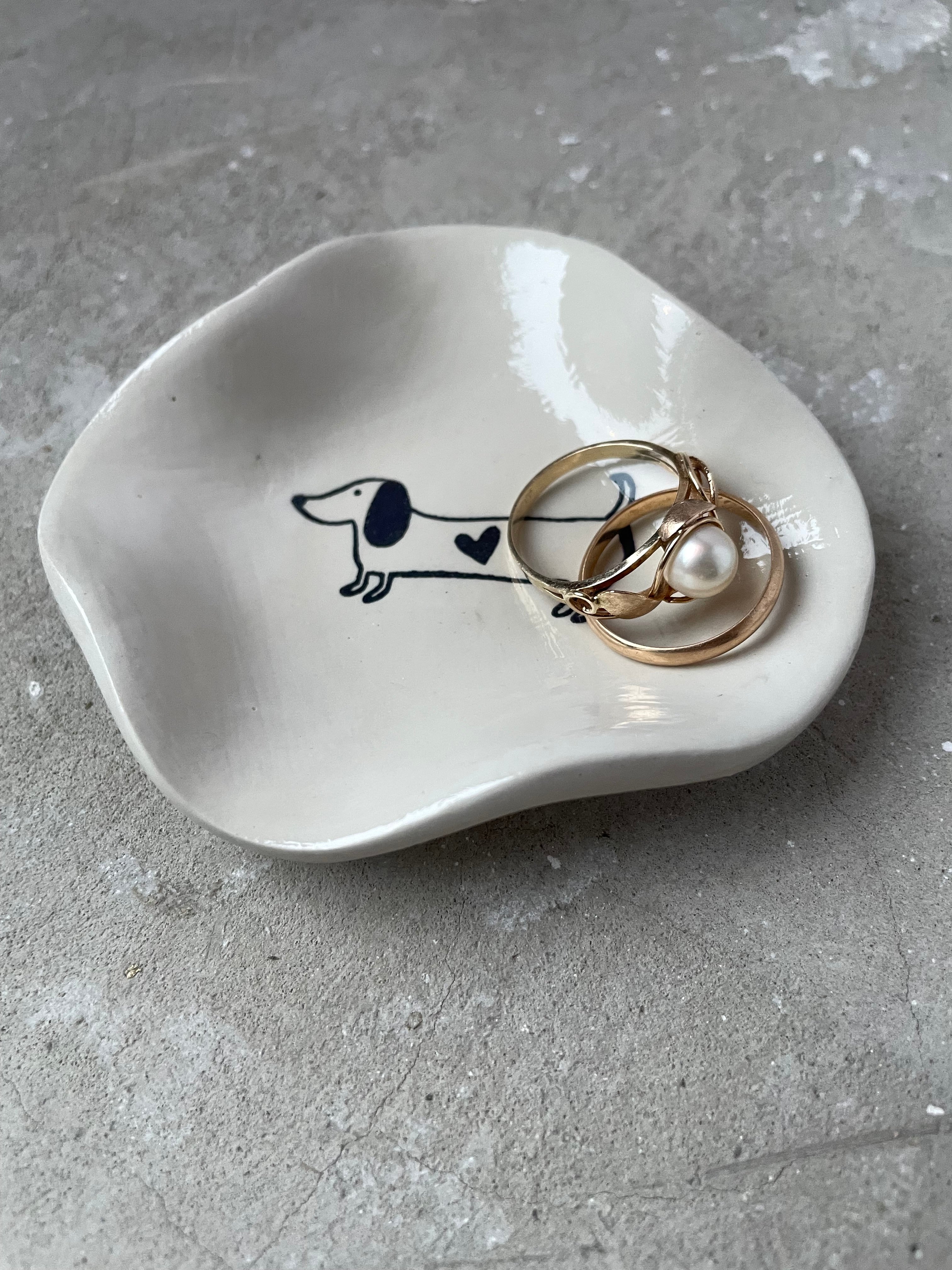 Modernes Keramikschälchen in Pantone-Beige, gegossen und mit glänzendem Finish versiegelt. Zeigt einen minimalistischen Dackel mit Herz – charmant, humorvoll und ideal für Hundemenschen. Perfekte Größe für Schmuck, Ringe, Schlüssel oder Kopfhörer. Handmade aus hochwertiger Gießkeramik, glatt, stabil und langlebig. Ideal als Geschenkidee für Dog Lovers, Dackelfans oder Freunde moderner, heller Wohnaccessoires. Leicht zu reinigen und vielseitig einsetzbar.