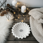 Große runde Rillenschale aus weißer Gießkeramik, etwa 20 cm Durchmesser. Die Schale hat eine charakteristische, wellenartige Rillenstruktur und in der Mitte ein winterliches Lettering in Schwarz. Fotografiert auf rustikalem Holztisch mit Kerzen, Tannenzweigen und warmen Lichtern im Hintergrund. Modernes, scandi-inspiriertes Design, ideal als winterliches Centerpiece, Kerzenschale oder Dekoteller. Handmade mit kleinen Materialnuancen.