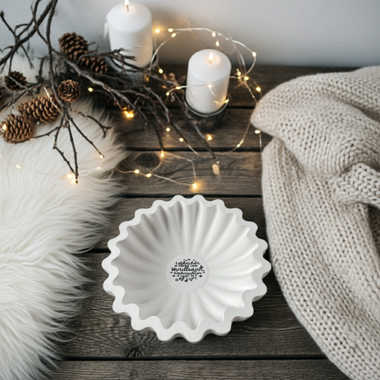 Große runde Rillenschale aus weißer Gießkeramik, etwa 20 cm Durchmesser. Die Schale hat eine charakteristische, wellenartige Rillenstruktur und in der Mitte ein winterliches Lettering in Schwarz. Fotografiert auf rustikalem Holztisch mit Kerzen, Tannenzweigen und warmen Lichtern im Hintergrund. Modernes, scandi-inspiriertes Design, ideal als winterliches Centerpiece, Kerzenschale oder Dekoteller. Handmade mit kleinen Materialnuancen.