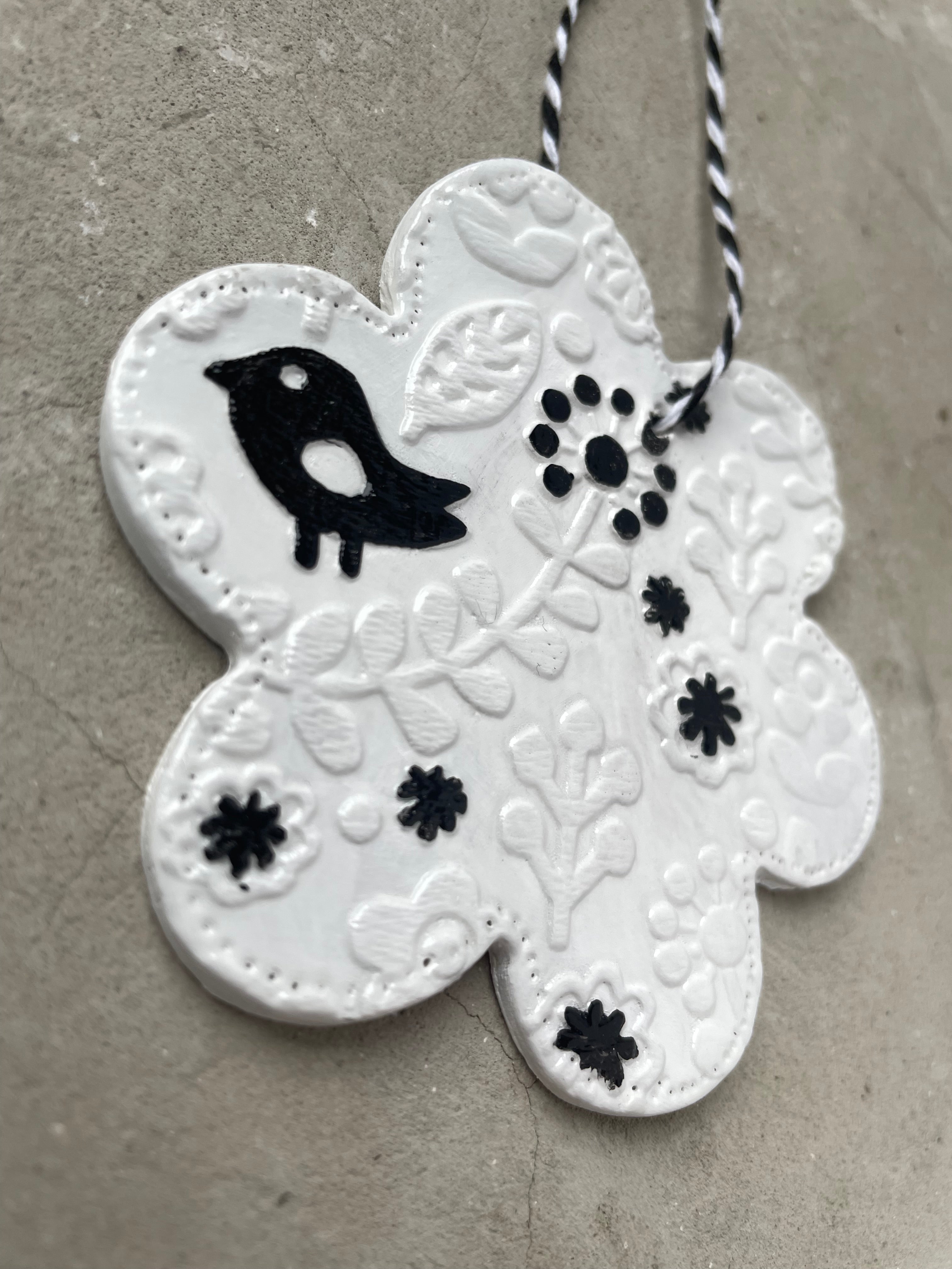 Anhänger in Blütenform aus weißer Keramik mit einem reliefartigen Blumenmuster und einer kleinen schwarzen Vogelfigur