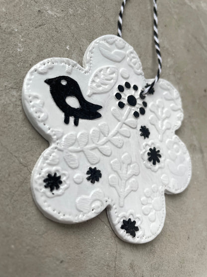 Anhänger in Blütenform aus weißer Keramik mit einem reliefartigen Blumenmuster und einer kleinen schwarzen Vogelfigur