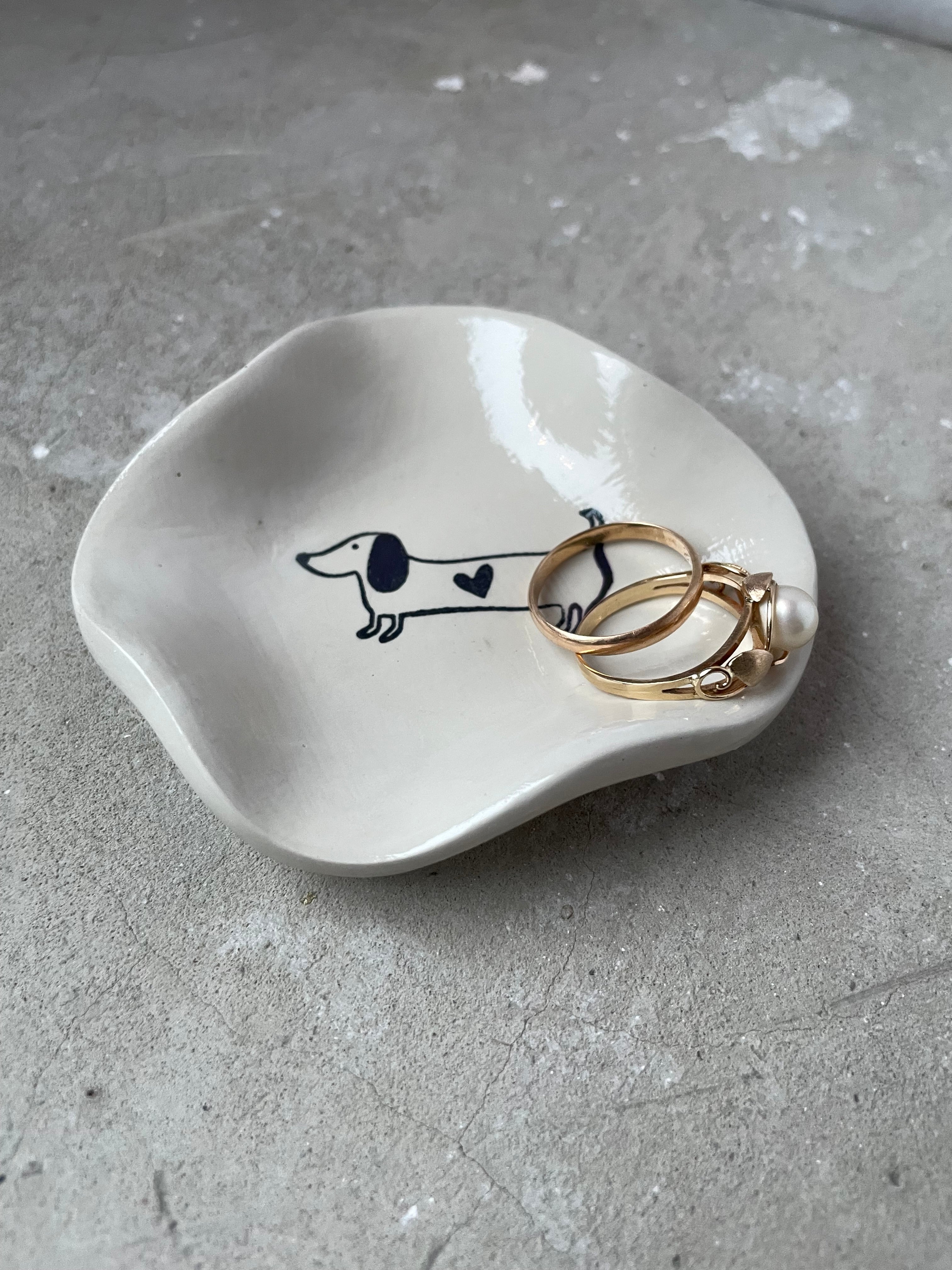 Kleines Dackel-Schälchen aus Keramikguss in Pantone-Beige mit glänzendem High-Gloss-Finish. Ideal als Ablage für Schmuck, Schlüssel oder kleine Alltagsdinge. Innen ein süßes Dackelmotiv mit Herz – perfekt für Hundeliebhaber, Tierfreunde und Fans von minimalistischer, moderner Dekoration. Jedes Stück wird einzeln gegossen, geschliffen und von Hand veredelt. Schlichte Form, glatte Oberfläche, handmade Charakter. Perfekt als Geschenk oder kleine Aufmerksamkeit.